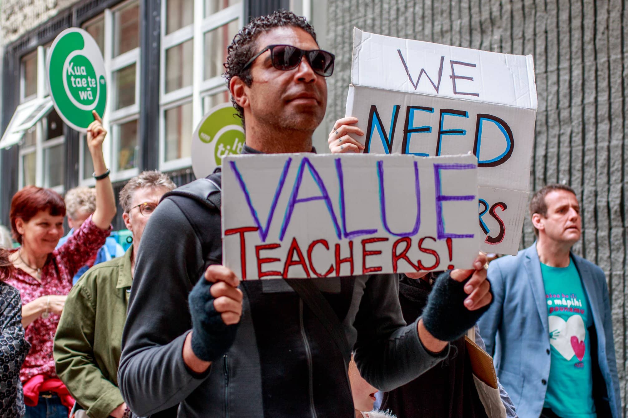 People march down the streets of Wellington, and we see the close up of several dedicated volunteers or teachers raising signs, "Value teachers", and "We need teachers"
