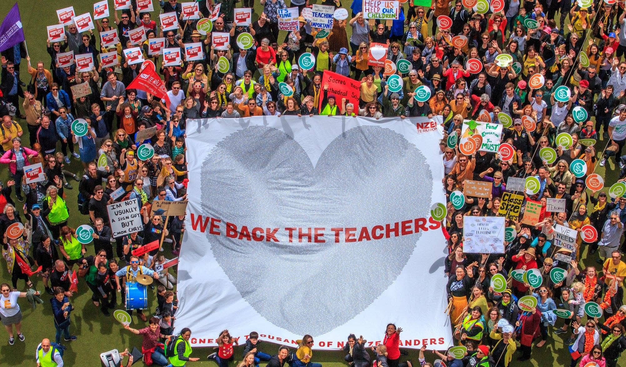 Huge and colorful crowd of union and teacher supporters in a field with a banner reading "We back the teachers"